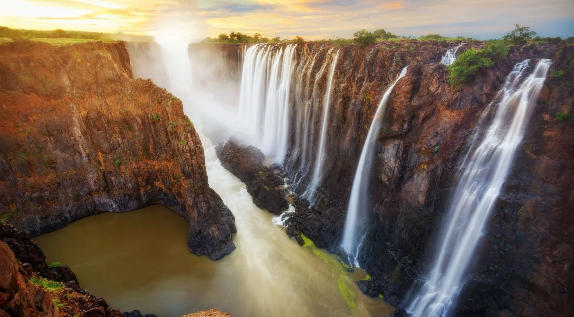 Victoria Falls gorge at golden hour, cascading water glowing in warm sunset light