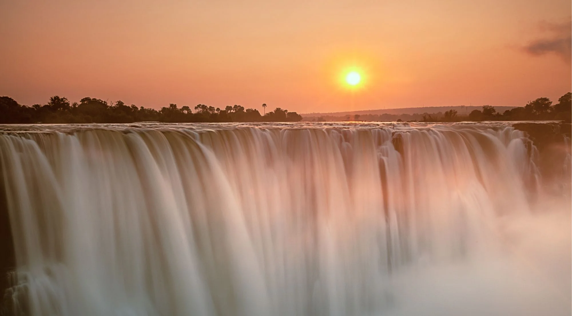 Victoria Falls full curtain of water at sunset, with an orange sun setting behind the African horizon and mist rising from the gorge