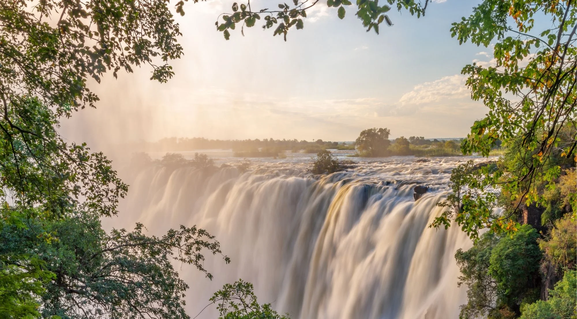 Victoria Falls framed by tropical foliage with sunlight breaking through the mist and spray, golden afternoon light
