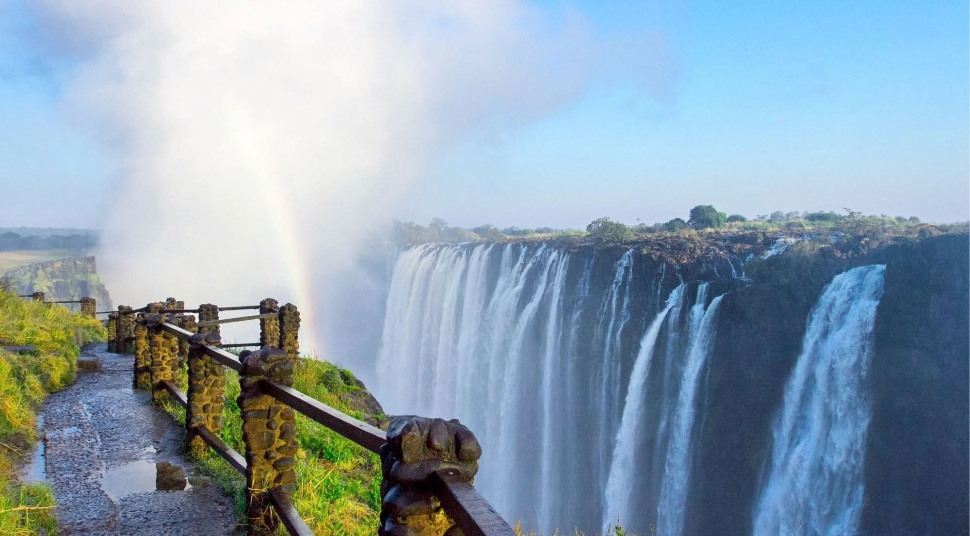 Rainbow forming over Victoria Falls seen from the national park walkway, with the full curtain of water and towering spray cloud