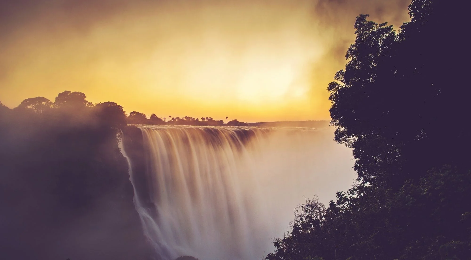 Victoria Falls at dramatic dusk with golden and purple sky, mist rising from the gorge and silhouetted trees in the foreground